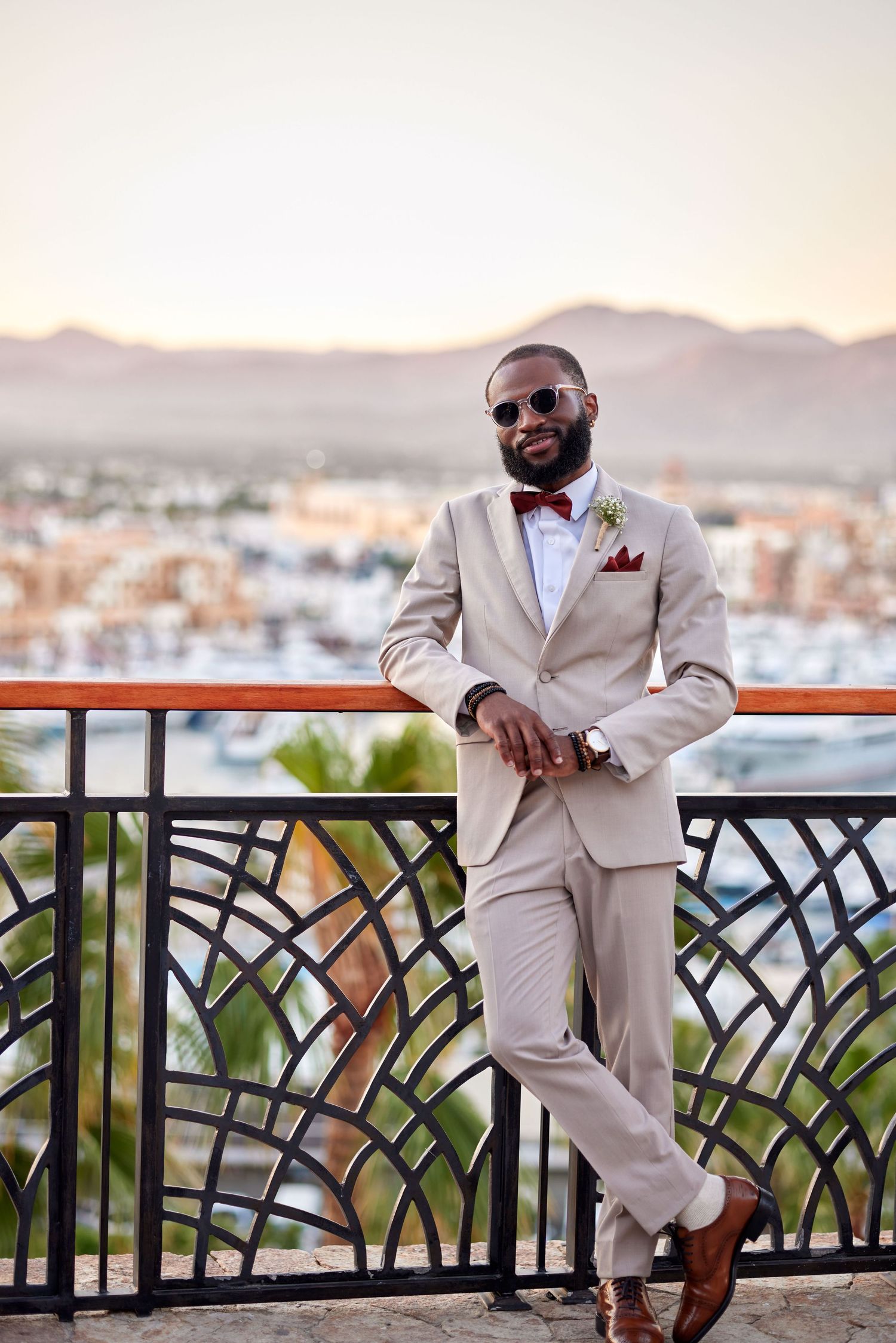 Stylish groom in beige suit with burgundy bow tie and sunglasses posing on terrace with mountain views.