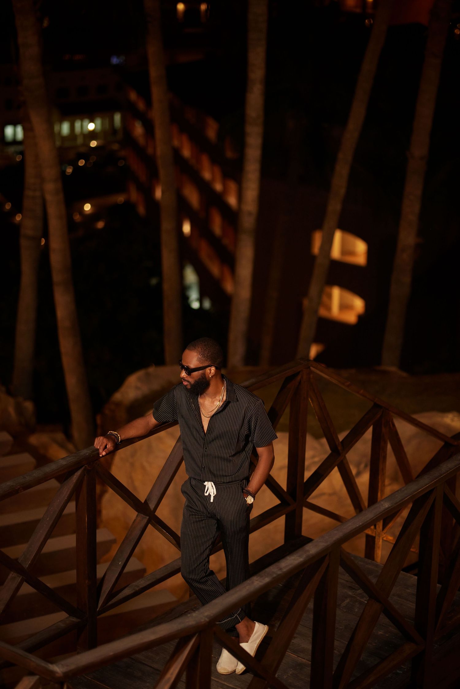 A stylish Black man in dark outfit and sunglasses stands on wooden stairs surrounded by palm trees at night.
