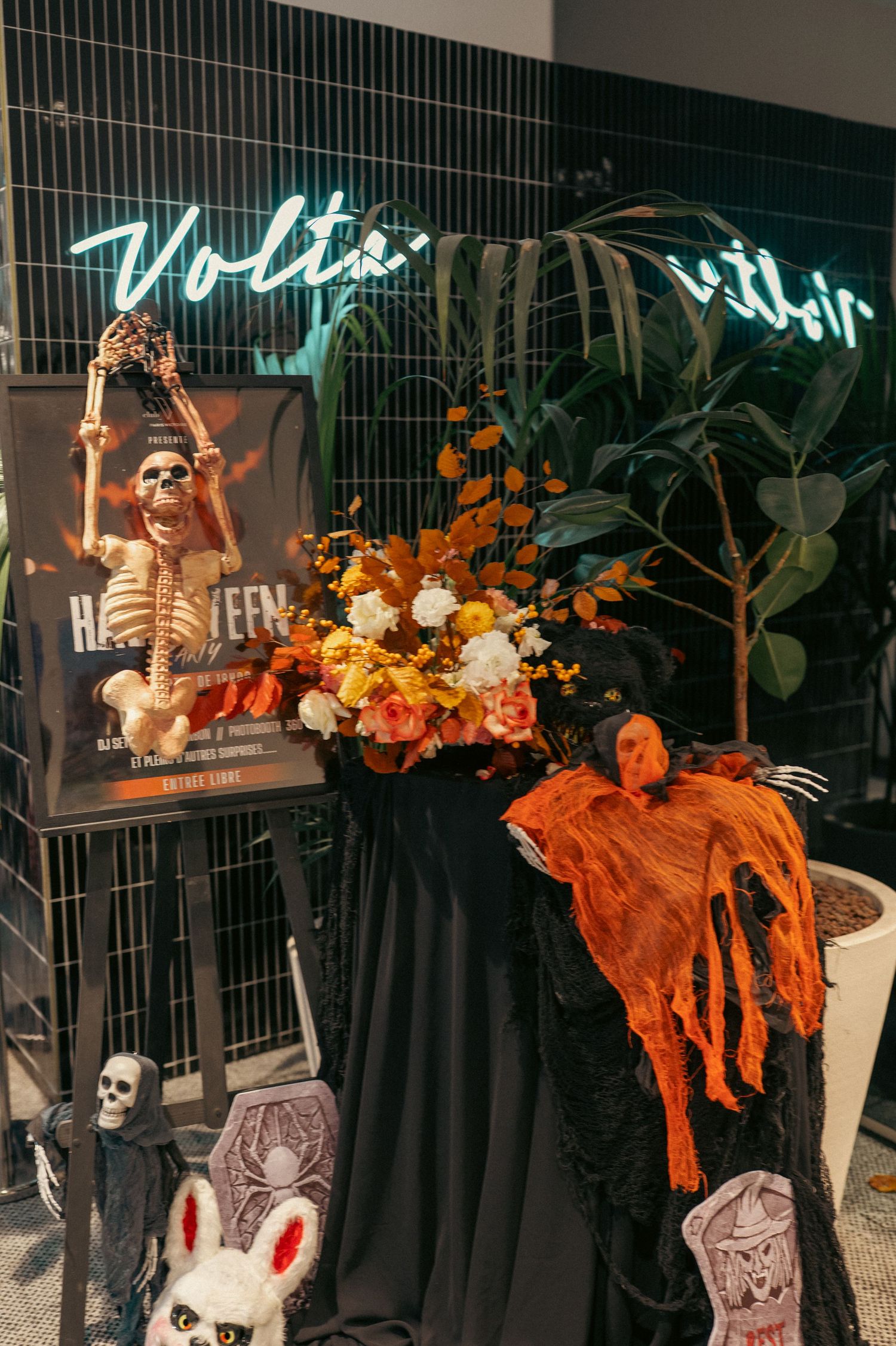 Affichage sur le thème d'Halloween avec squelette, arrangement floral orange et décorations effrayantes sous une enseigne au néon.
