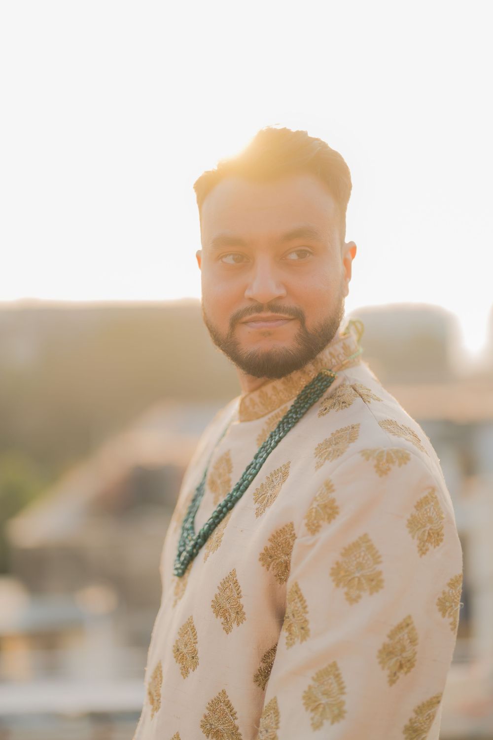 Man in traditional Indian sherwani with gold embroidery and green necklace poses outdoors in golden hour sunlight.