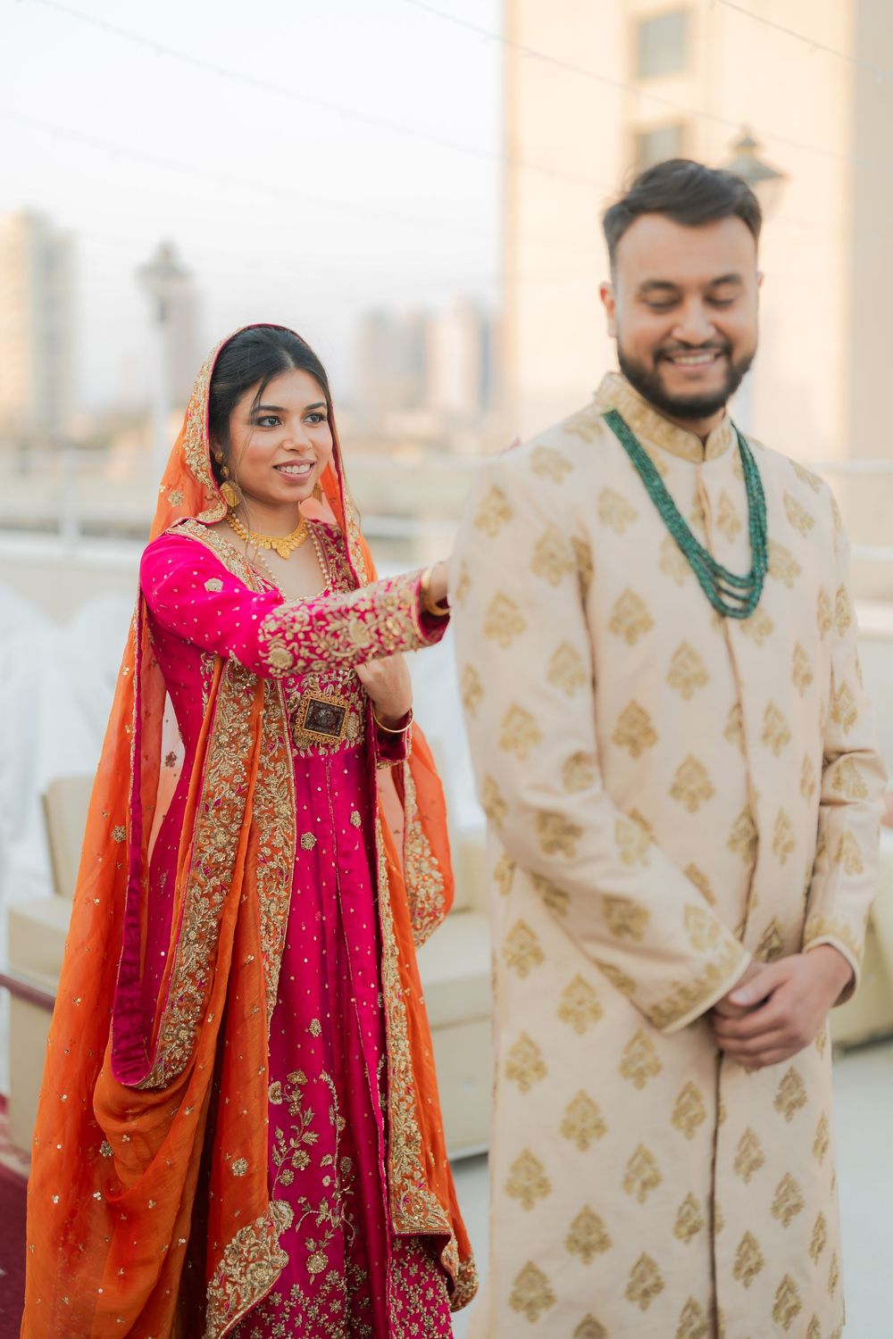 Indian bride in pink and orange lehenga holds groom's hand as he smiles in cream sherwani with green beaded necklace.