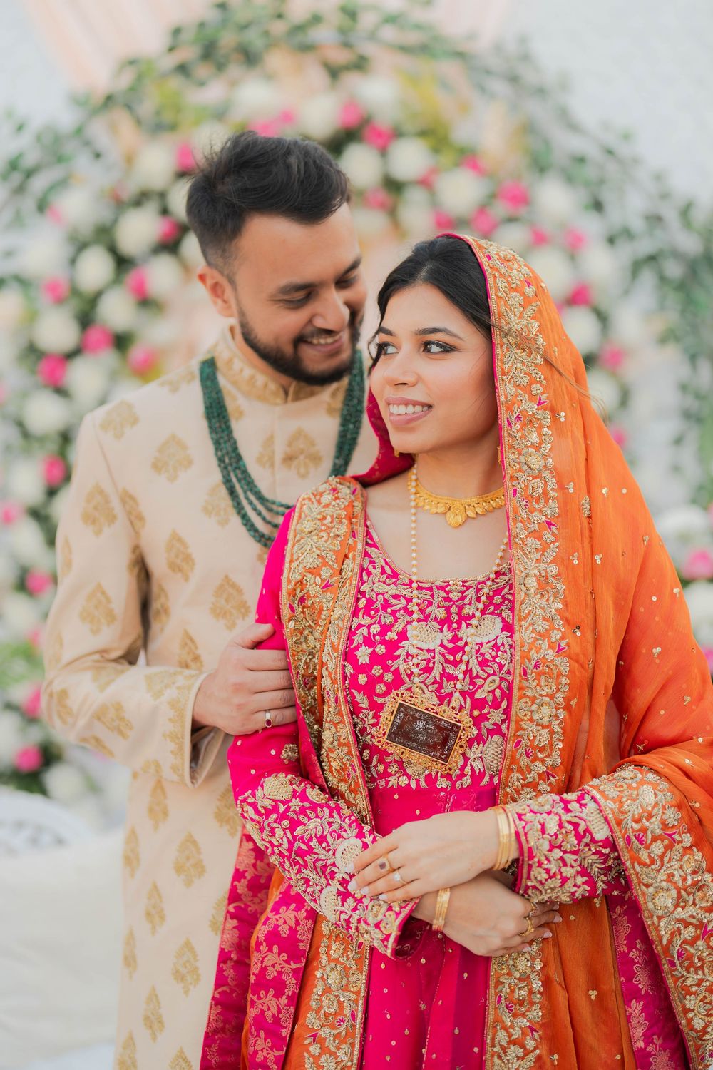 Couple in traditional South Asian wedding attire, bride in pink and orange lehenga with gold jewelry, groom in cream sherwani.