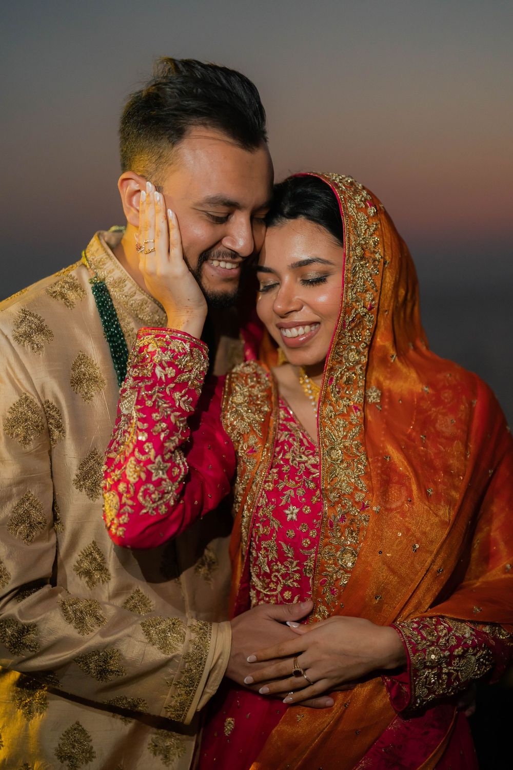 South Asian couple in traditional wedding attire sharing a tender moment at dusk, bride in red dupatta.