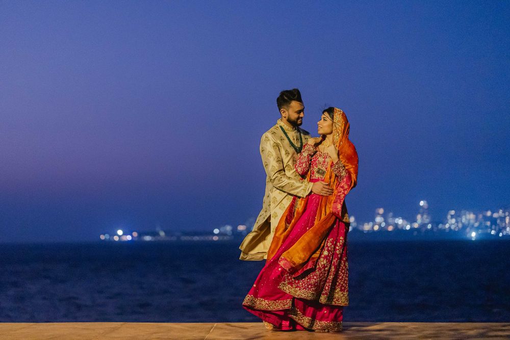 Couple in traditional Indian wedding attire embracing romantically by the sea at twilight with city lights backdrop.
