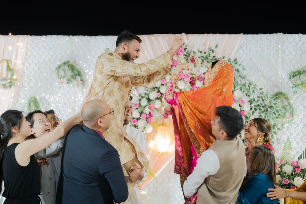 Groom in gold sherwani lifted by guests at vibrant Indian wedding ceremony with floral backdrop.