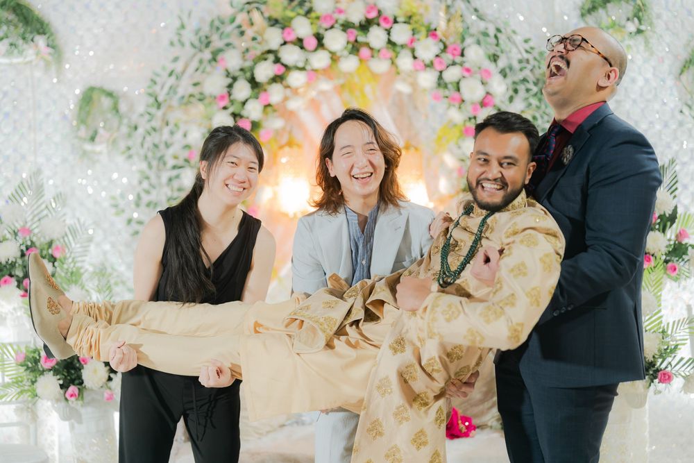 Four friends laugh joyfully at a floral wedding celebration, with two men lifting a woman in a festive golden outfit.
