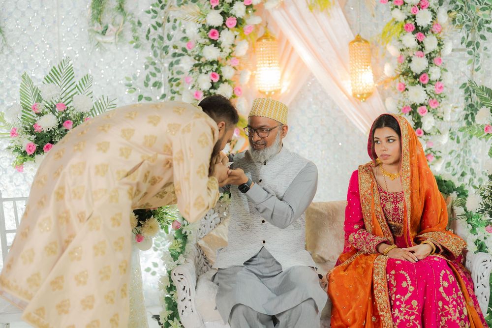A South Asian wedding ceremony with a bride in pink and groom in cream attire, blessed by an elder amid floral decorations.