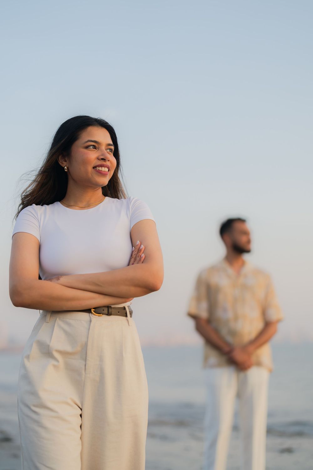 A smiling woman with crossed arms stands on a beach at sunset, with a man standing in the background.