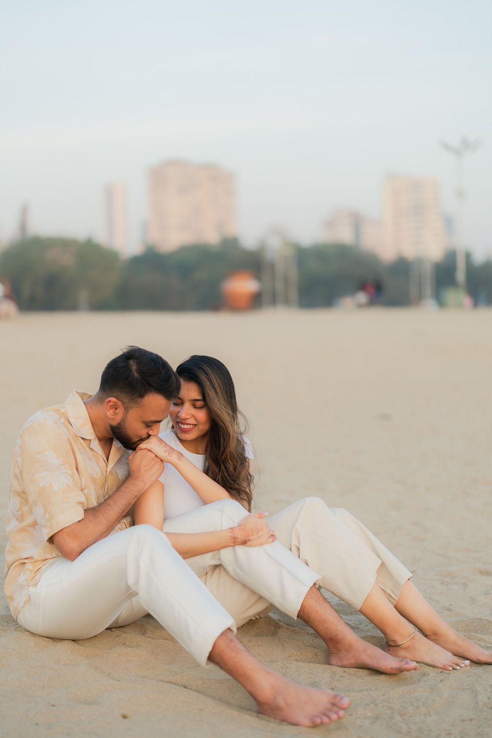 Romantic couple sitting barefoot on sandy beach at sunset, with city skyline in soft focus background.