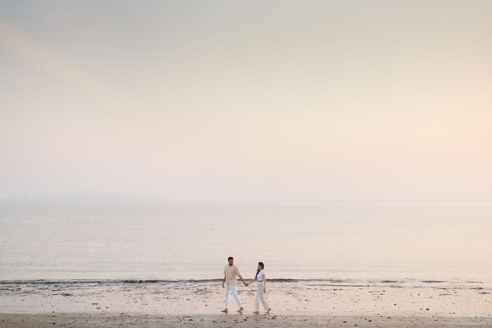 Couple in white holding hands walking on a serene beach at sunset with calm ocean waters in the background.
