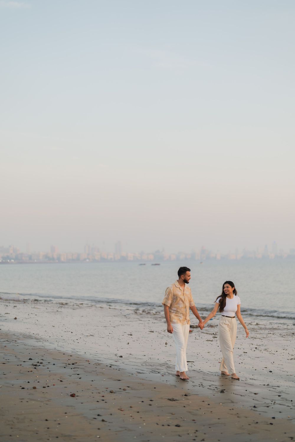 A couple walks hand-in-hand along a serene beach at dusk, with a city skyline visible across the calm water.