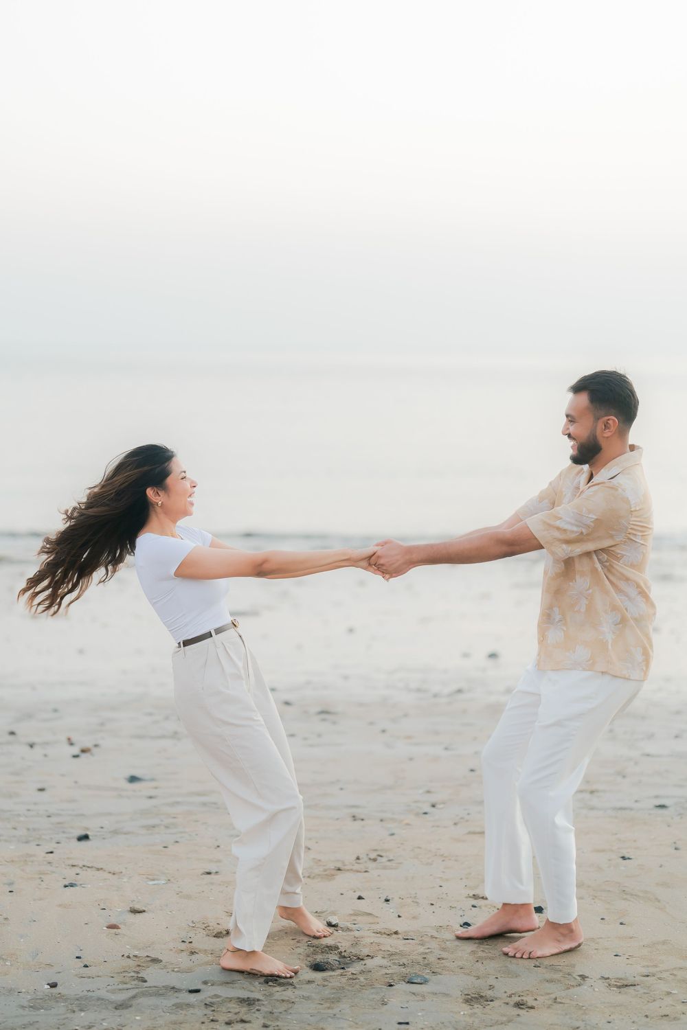 A couple holds hands and dances joyfully on a sandy beach at sunset, dressed in casual white and beige outfits.
