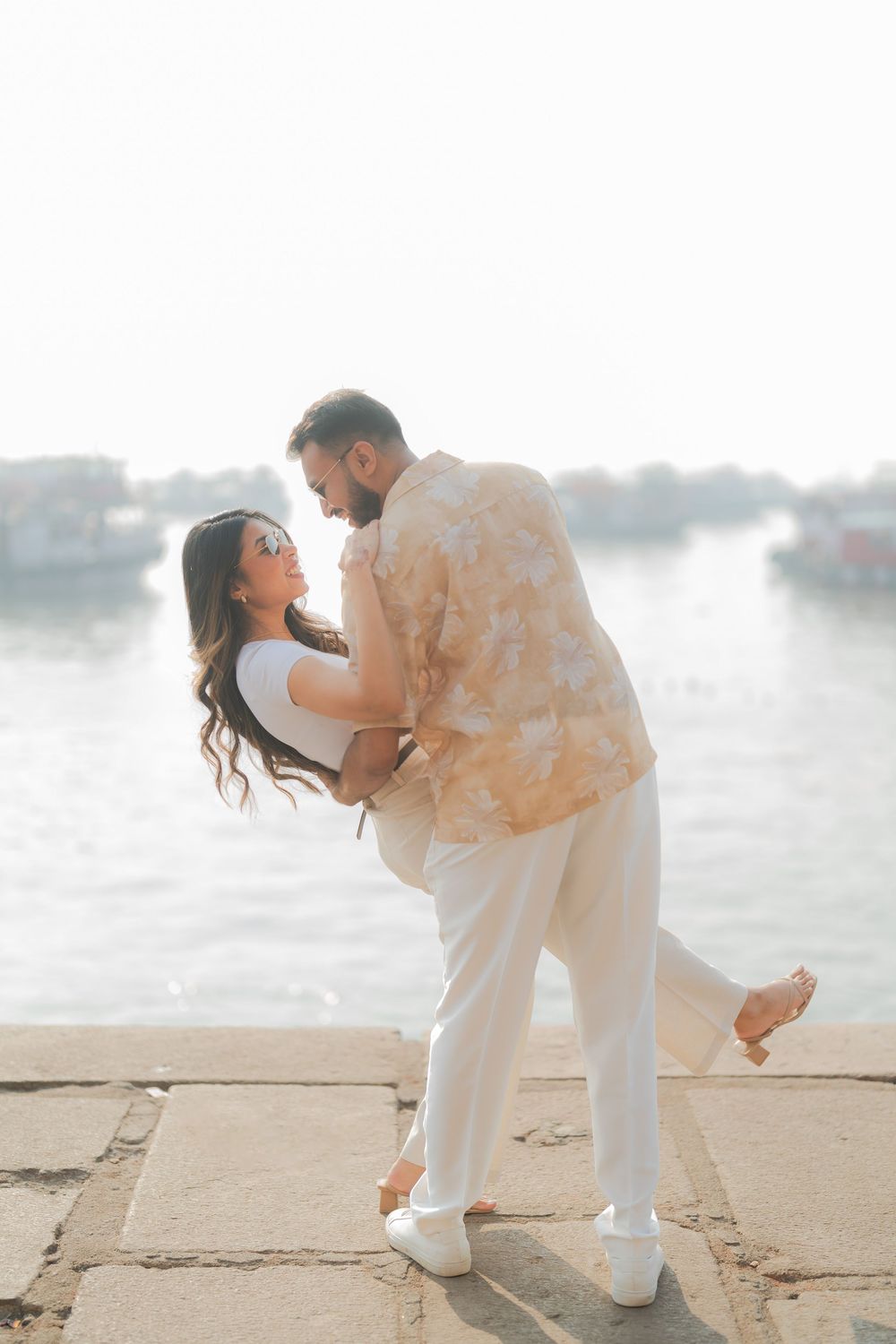 Couple dancing romantically on a waterfront promenade, man dipping woman in white outfit, boats visible in background.