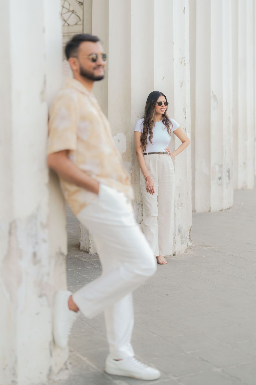 Stylish couple posing by white columns outdoors, man in beige shirt and white pants, woman in white outfit with sunglasses.