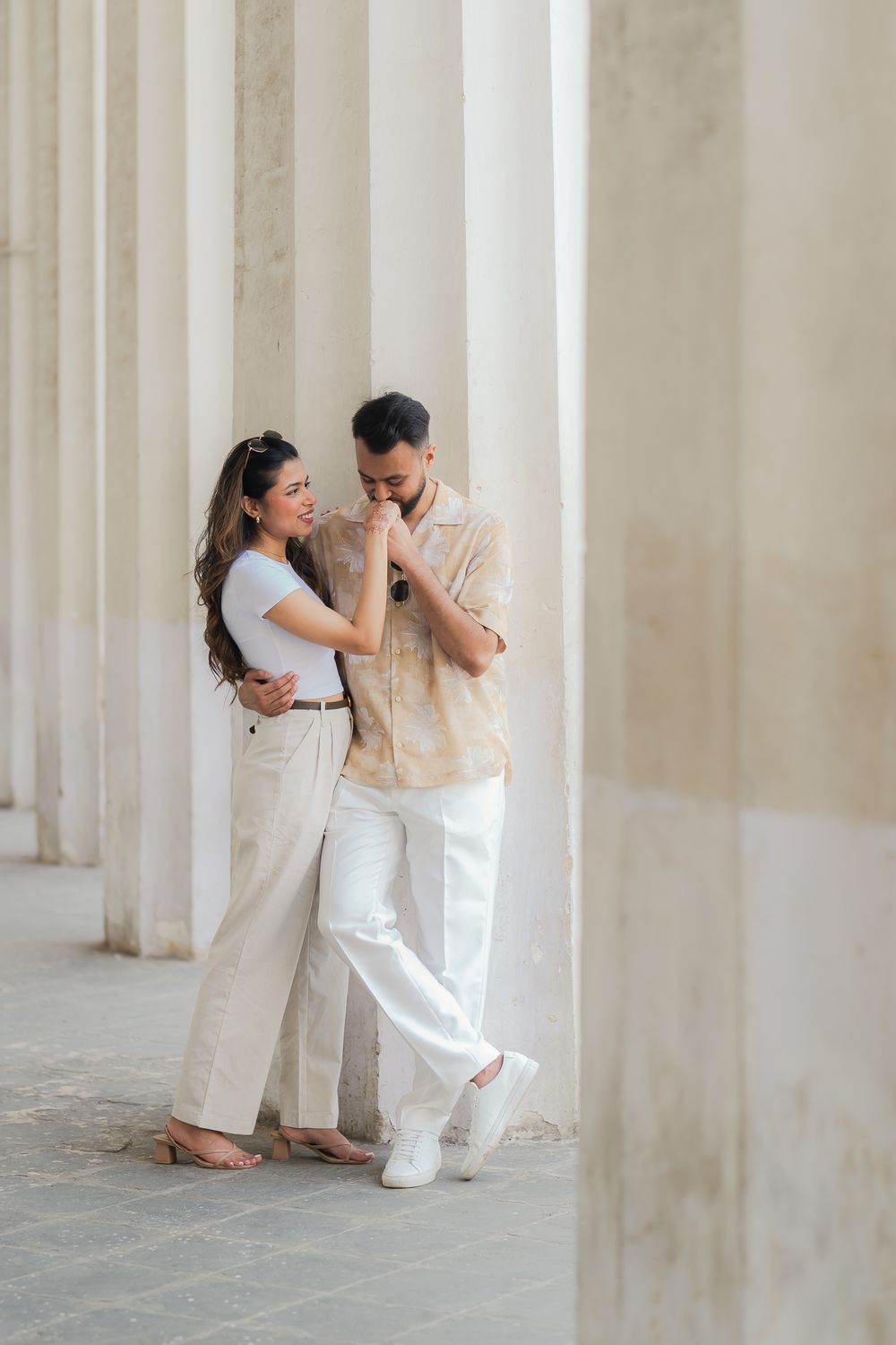 Romantic couple in neutral tones embracing between elegant white columns, man kissing woman's hand.