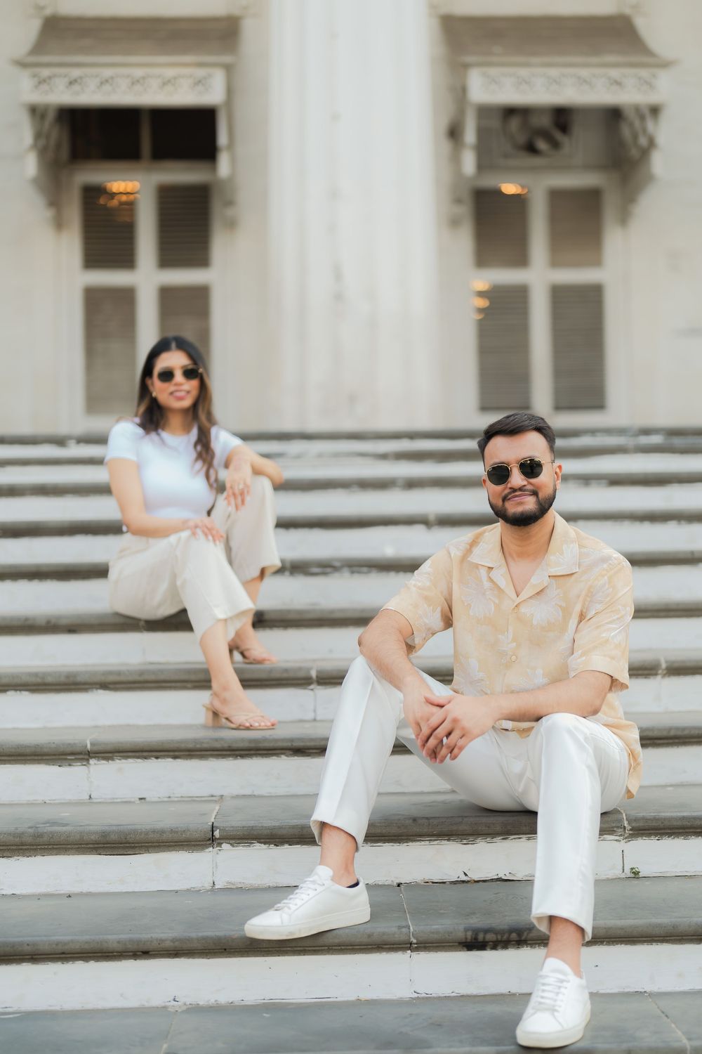 Stylish couple in beige outfits sitting on white marble steps of an elegant building, wearing sunglasses.
