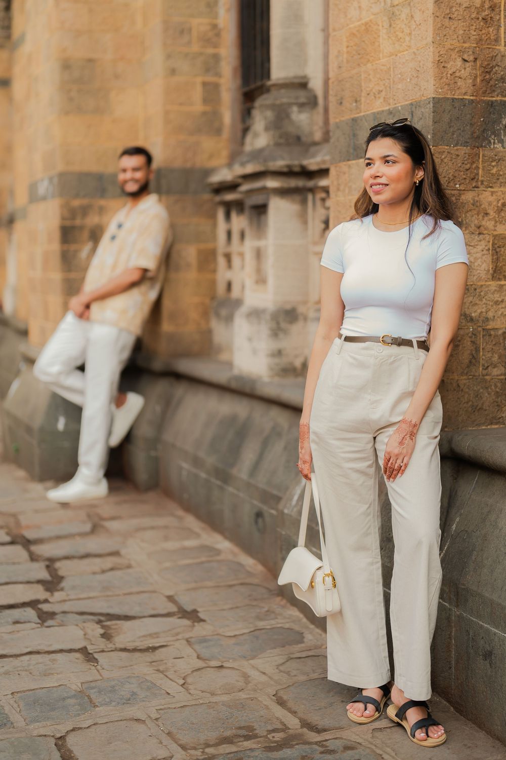 A stylish couple leaning against a historic stone building on a cobblestone street, dressed in neutral tones.