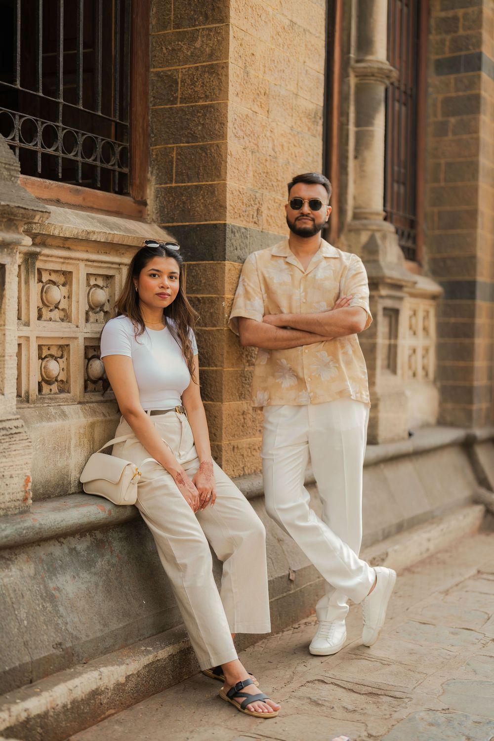 A stylish couple poses against an ornate historic building, wearing coordinated beige and white outfits in a urban setting.