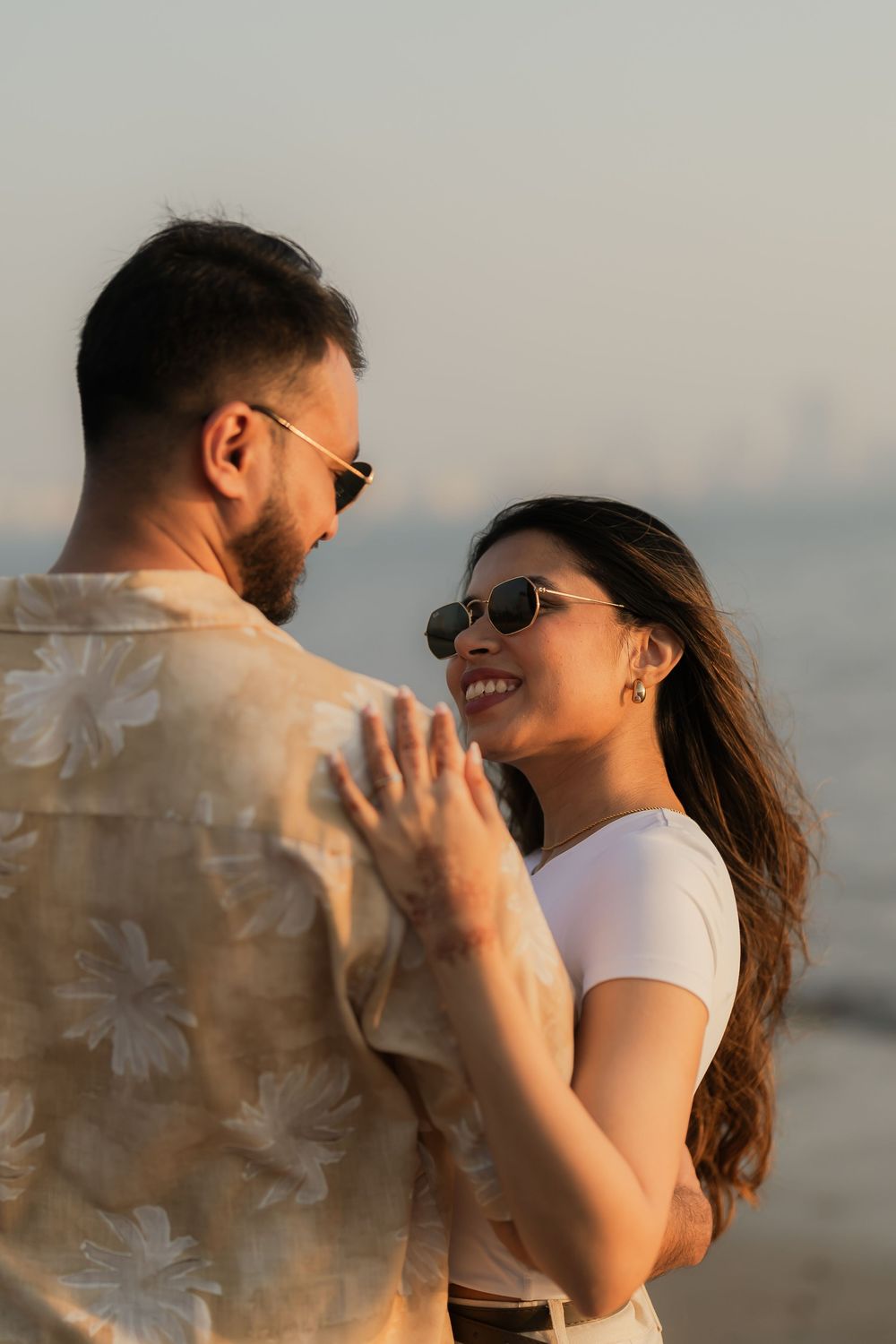 Couple in sunglasses sharing a romantic moment on a beach at sunset, woman smiling warmly at man in floral shirt.