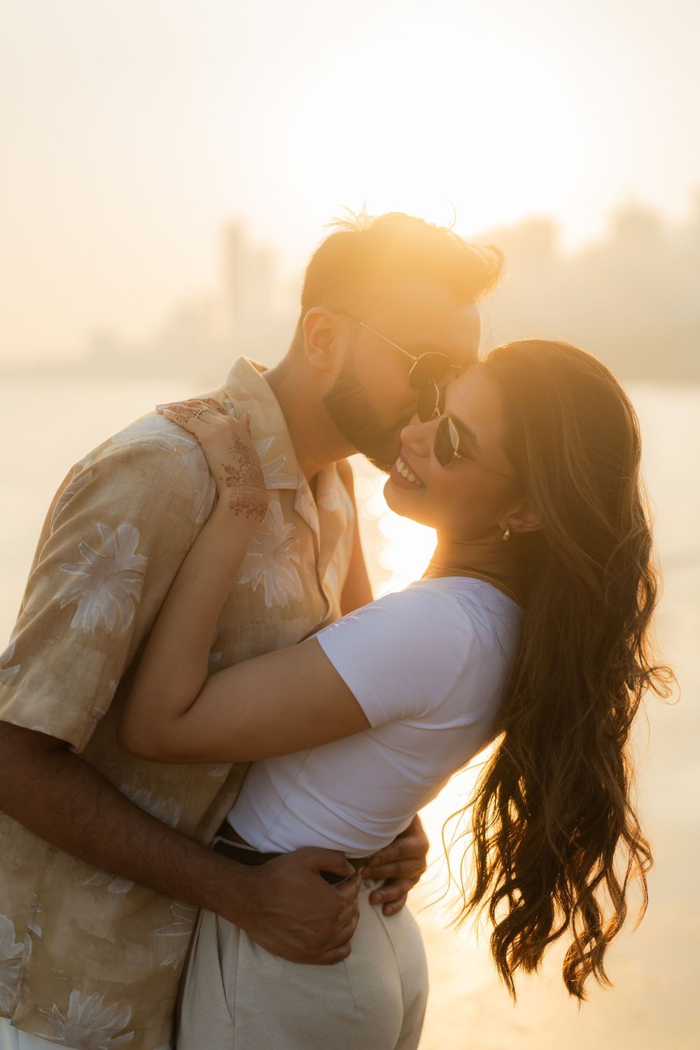 Couple embracing romantically at golden sunset by the waterfront, woman smiling in sunglasses, man kissing her forehead.