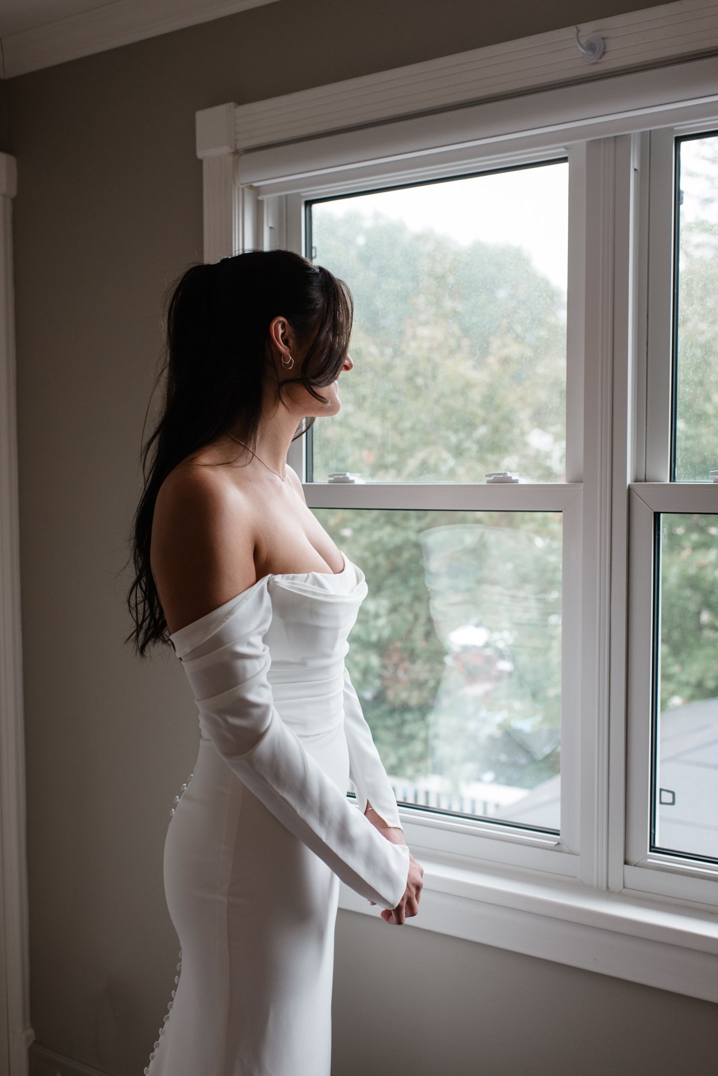 A figure in a white off-shoulder dress gazes out of a bright window in a minimalist room interior.