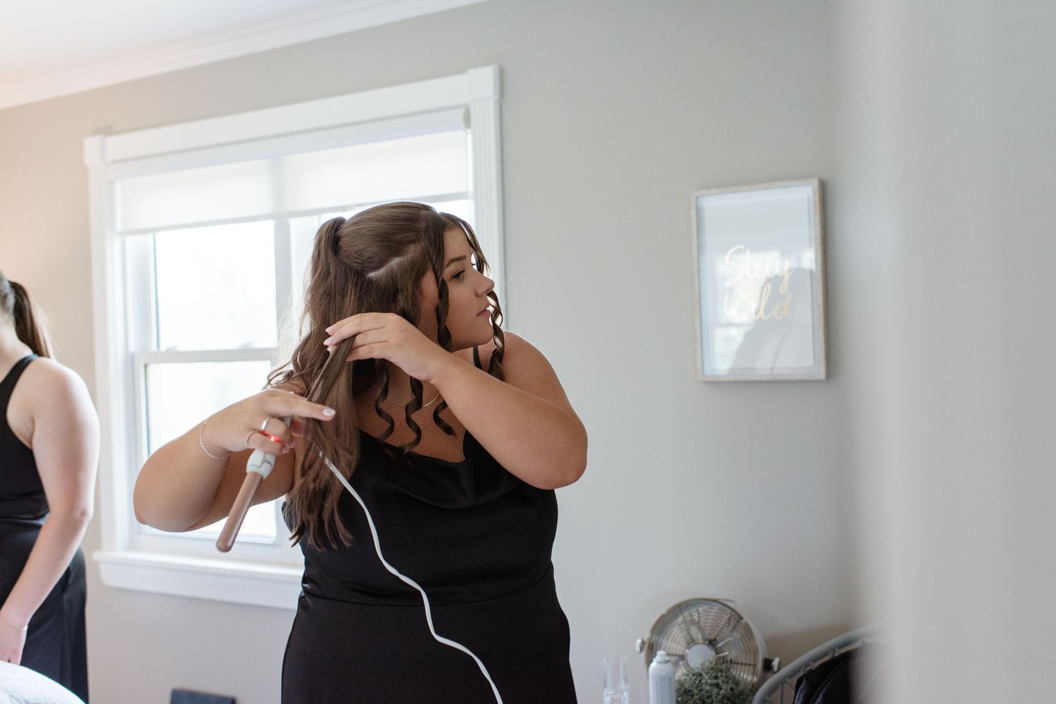 A hairstylist curling a woman's hair indoors near a bright window in a softly lit room.