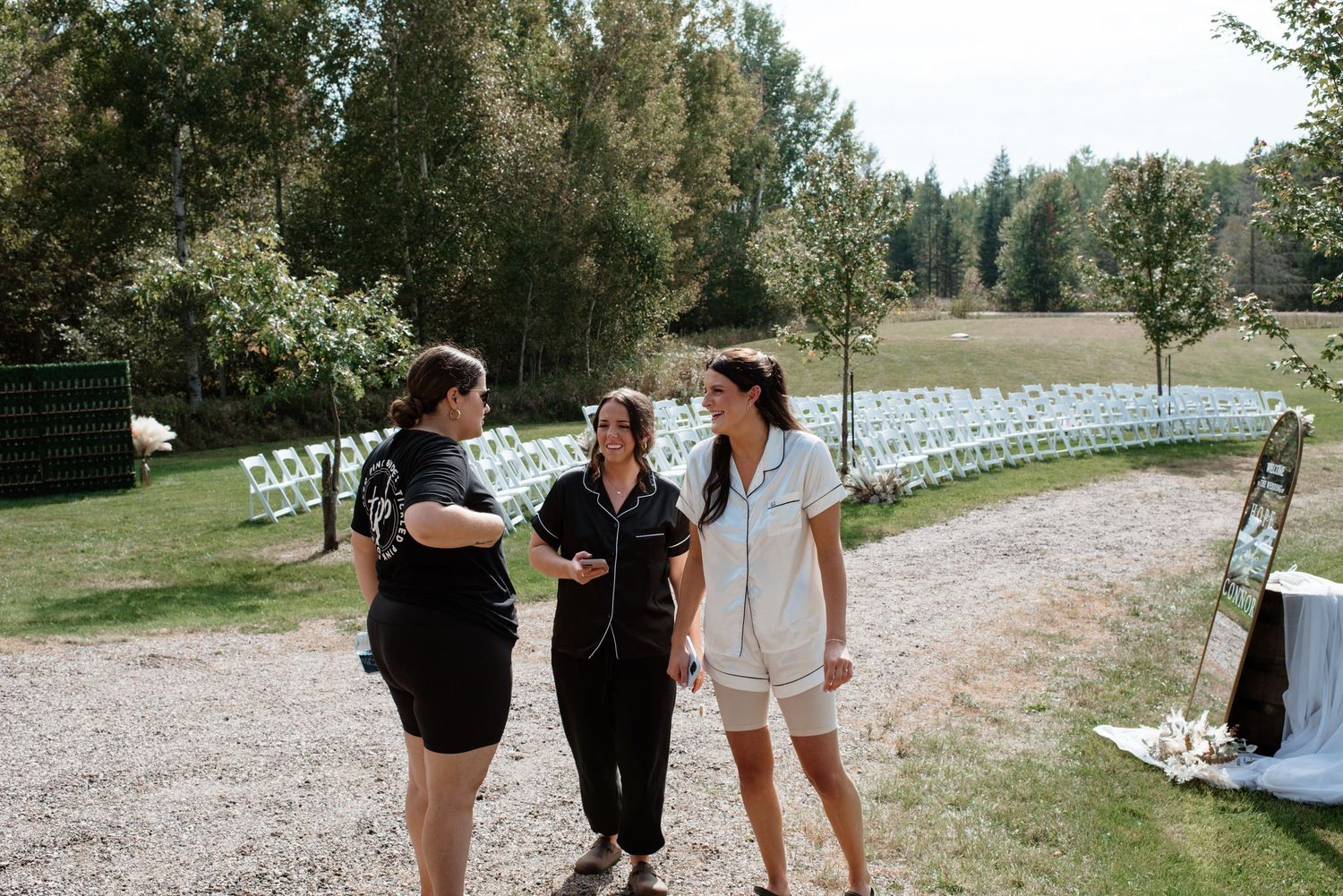 Three women conversing outdoors at a wedding venue with white chairs and greenery in background.