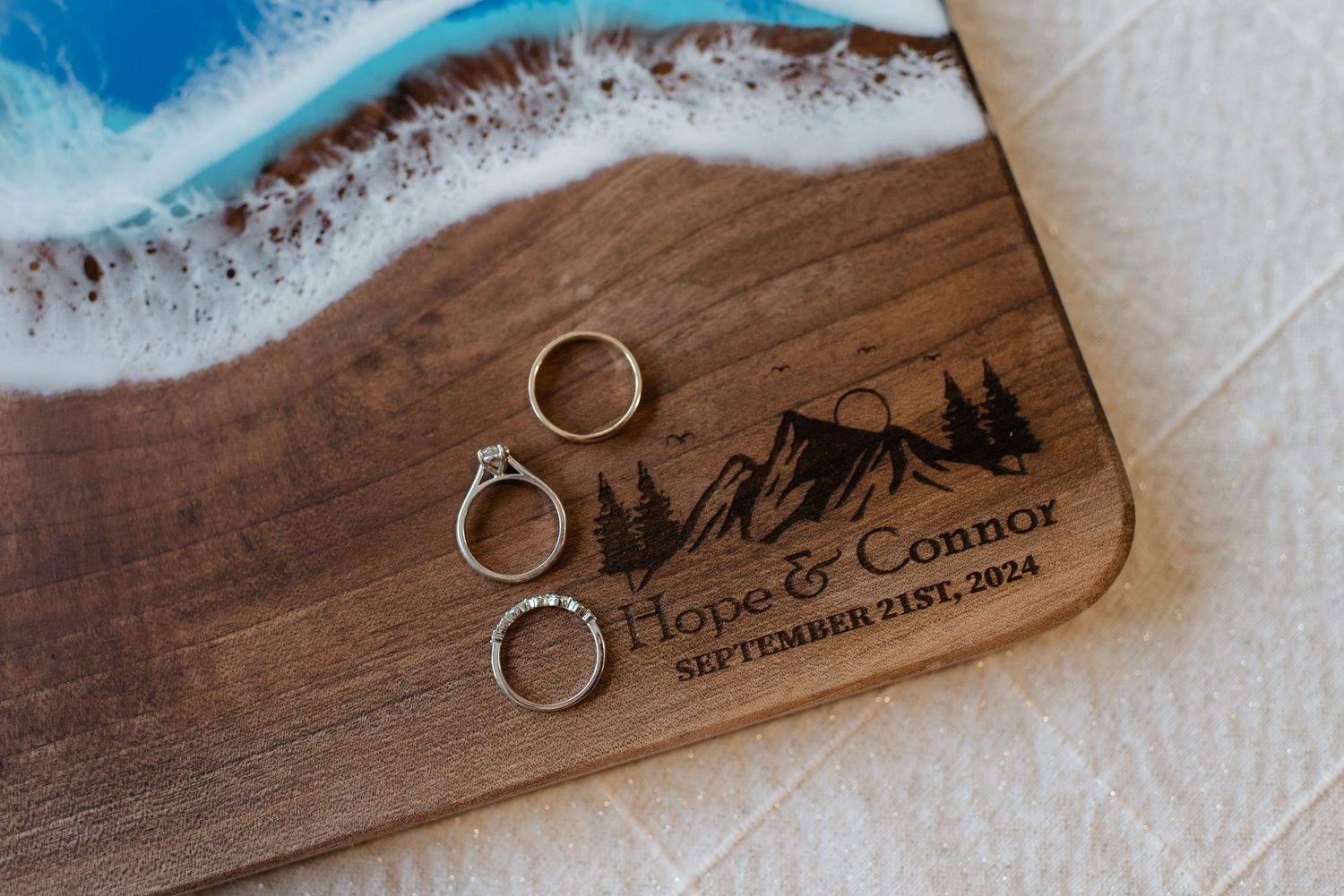 Wedding rings displayed on engraved wood board for Hope and Connor, September 21st 2024, with ocean resin art.