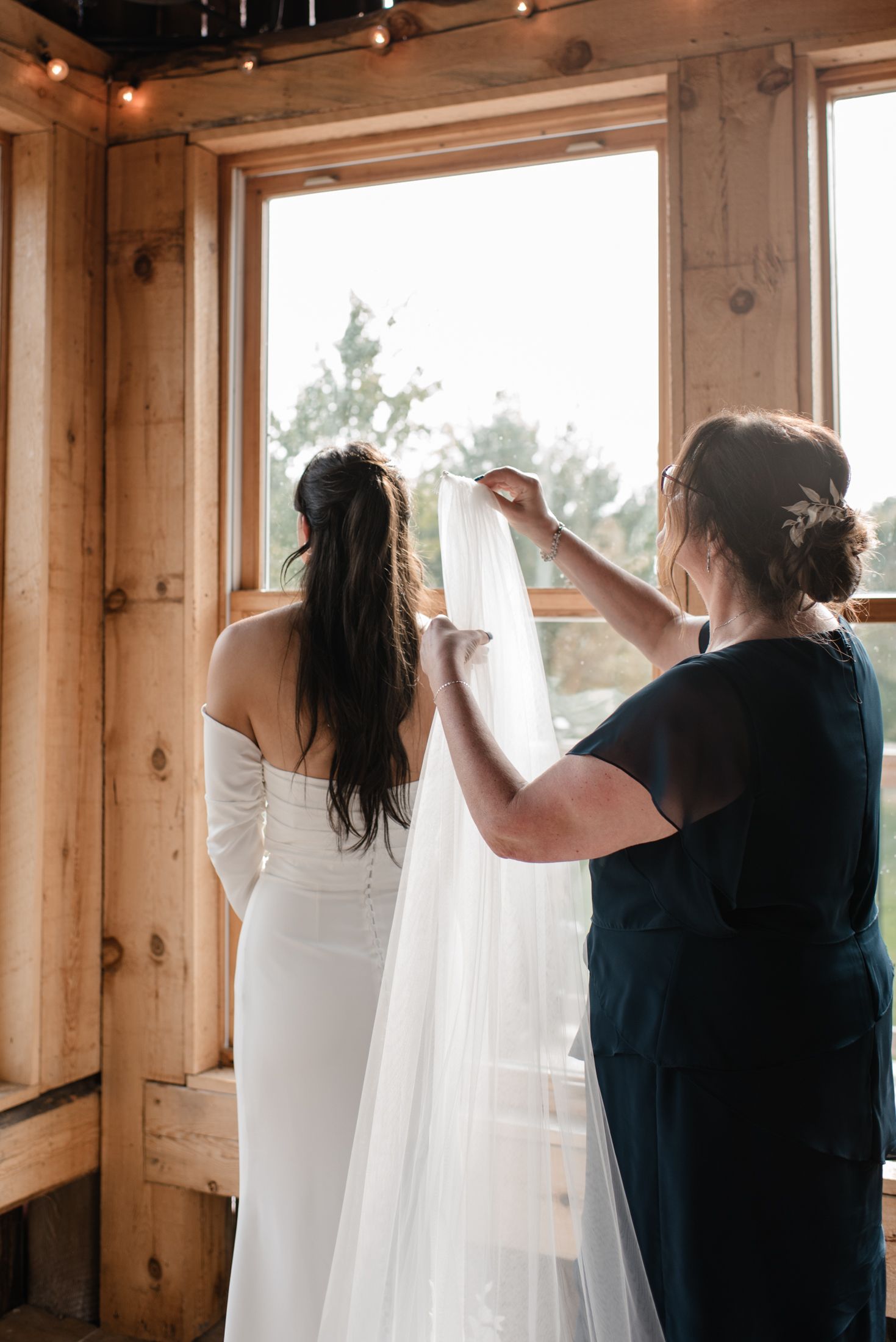 Bridesmaid helping bride with veil in rustic wooden venue near window on wedding day.