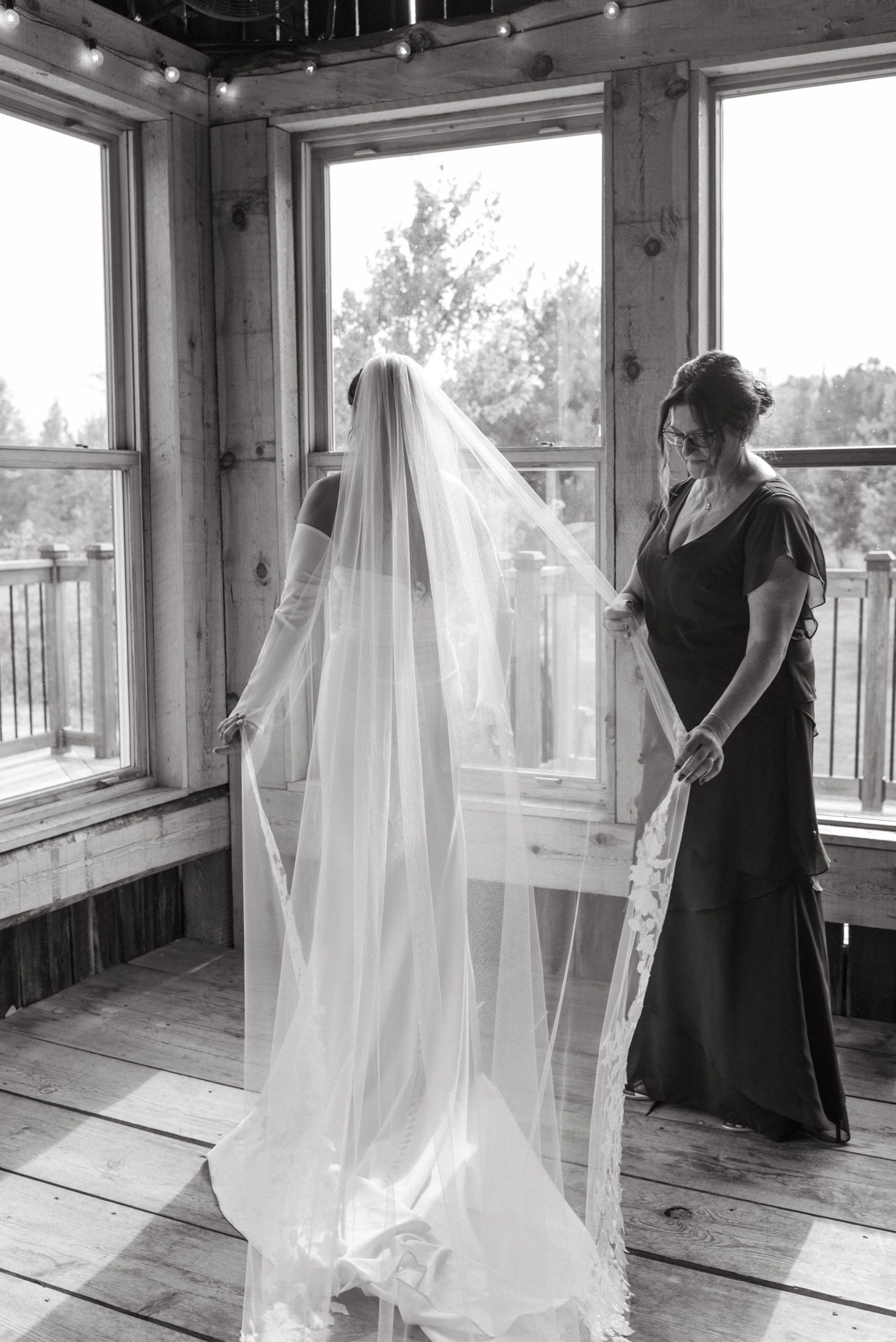 Black and white photo of a bride in a long veil being assisted by a bridesmaid in a rustic wooden venue.