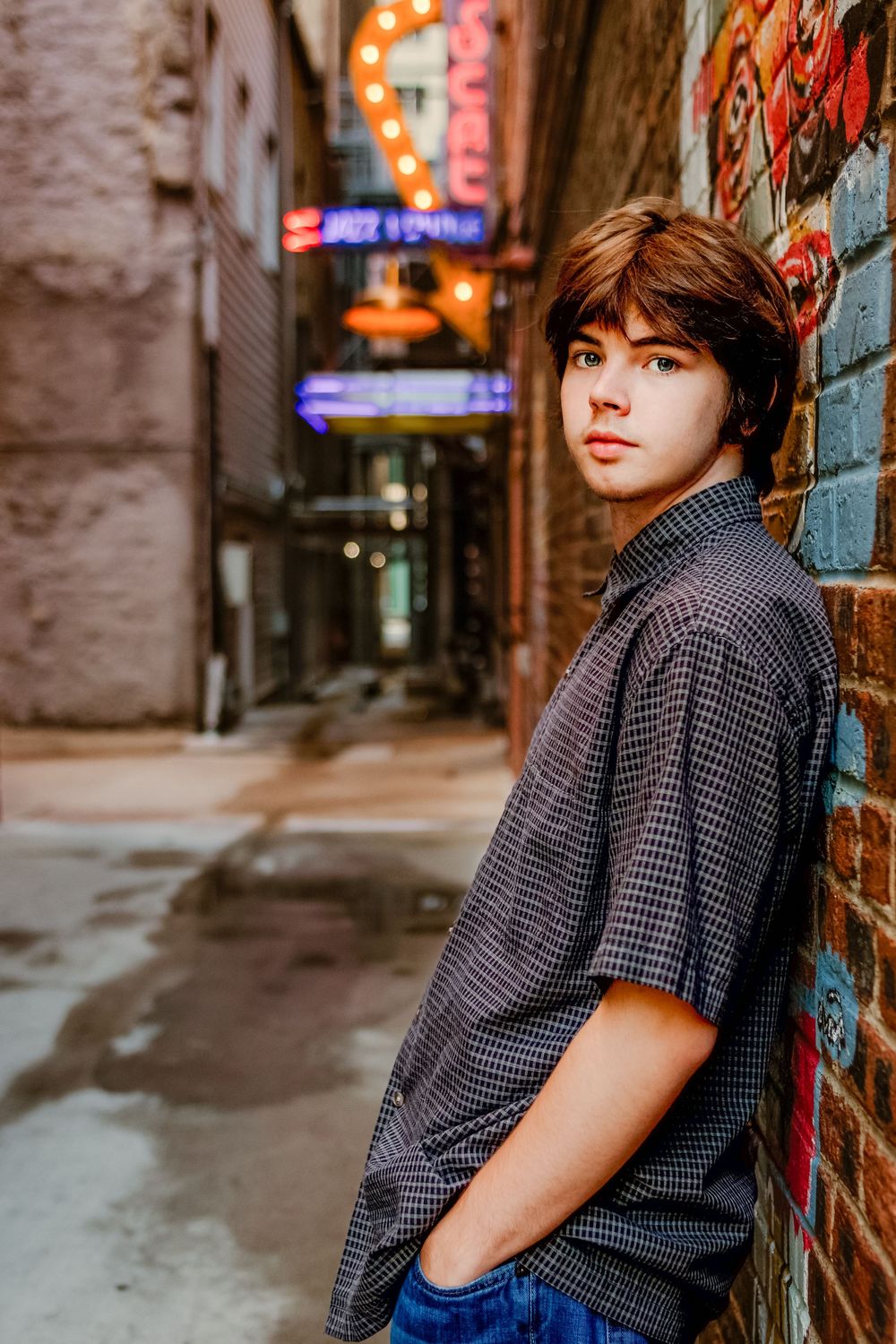 Teen boy leaning against graffiti brick wall in urban alley with neon signs in background.