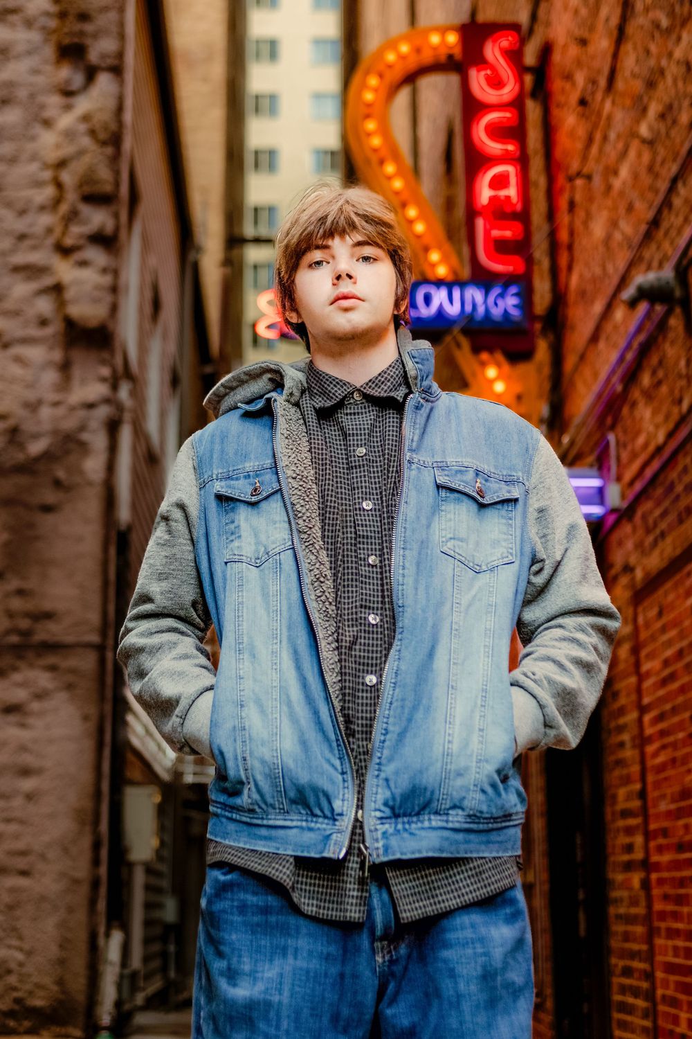 Young man in denim jacket poses in urban alley with neon Scat Lounge sign glowing behind him.