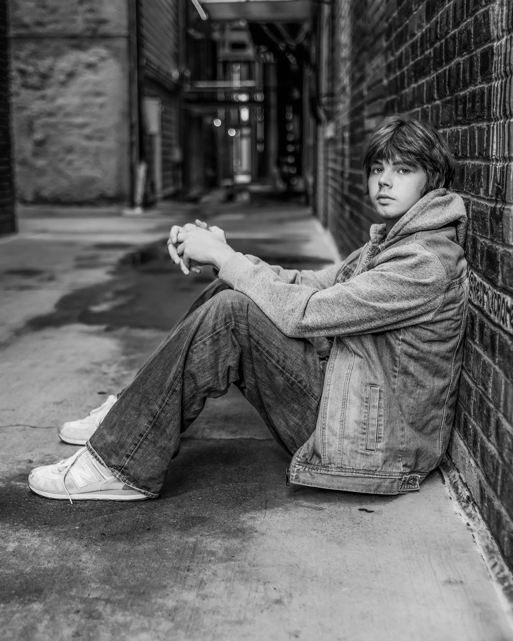 Black and white photo of a young person sitting against a brick wall in an urban alley, wearing jeans and a denim jacket.