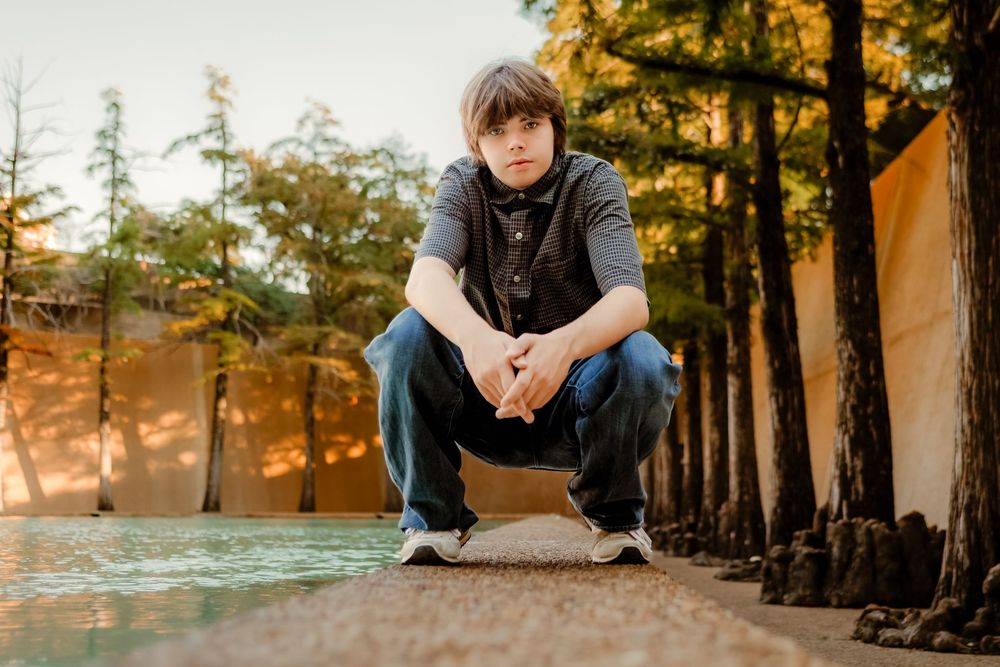 Young man crouching near a reflective pool surrounded by tall trees during golden hour outdoors.