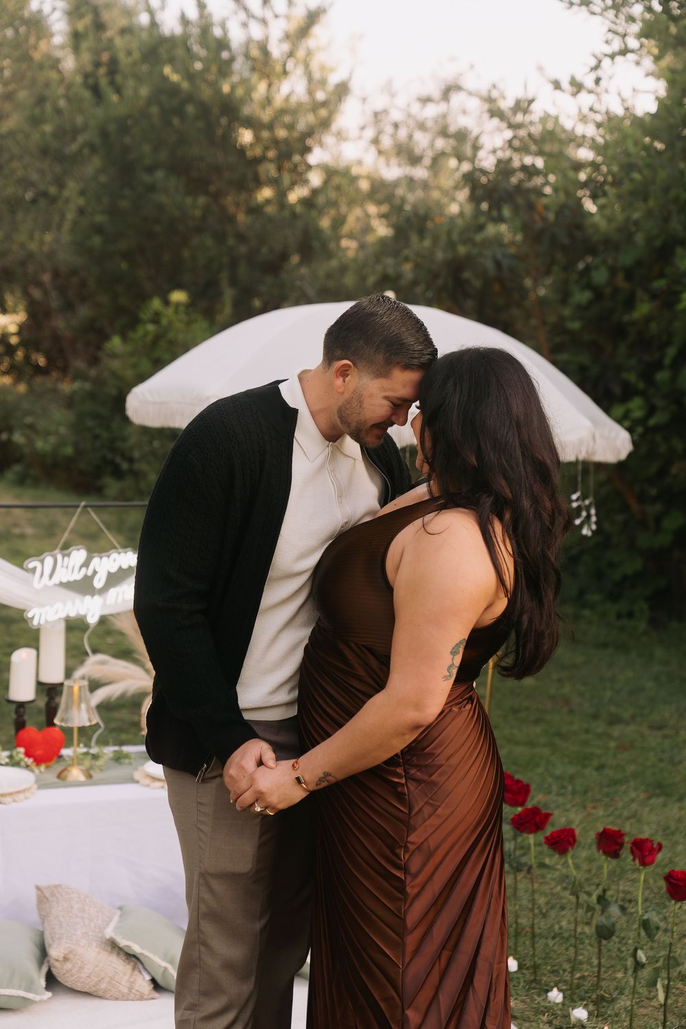Couple sharing a romantic moment at an outdoor proposal setup with roses, candles, and a will you marry me sign.