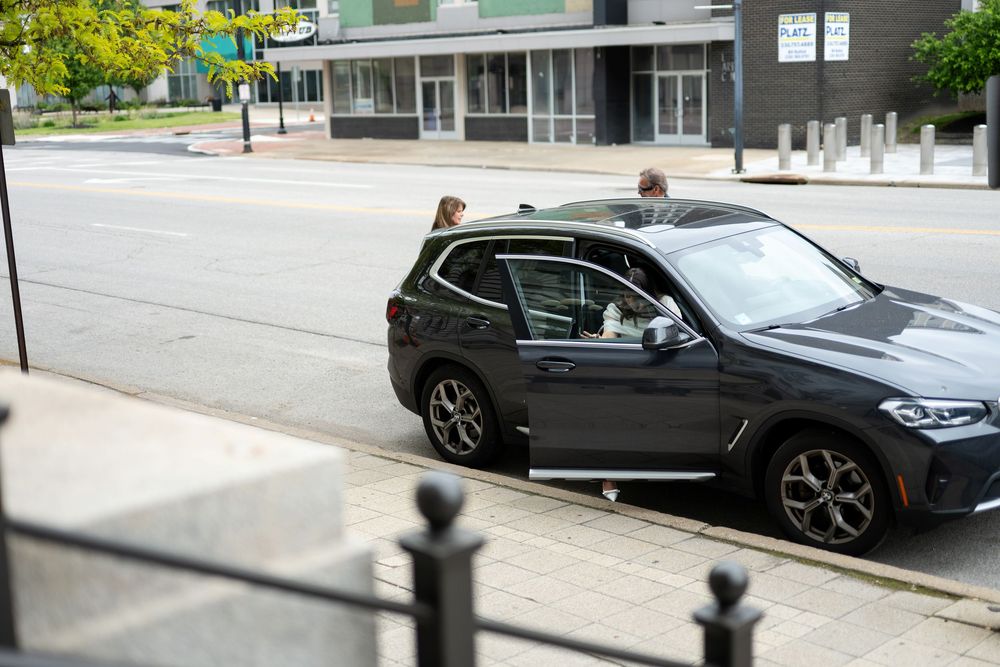 Dark BMW SUV parked on urban street curb with passenger entering vehicle in city setting.