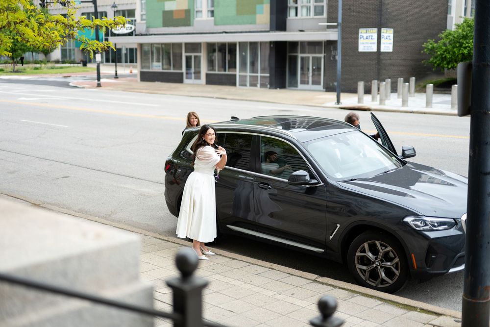 Bride in white dress stands beside dark SUV on urban city street.