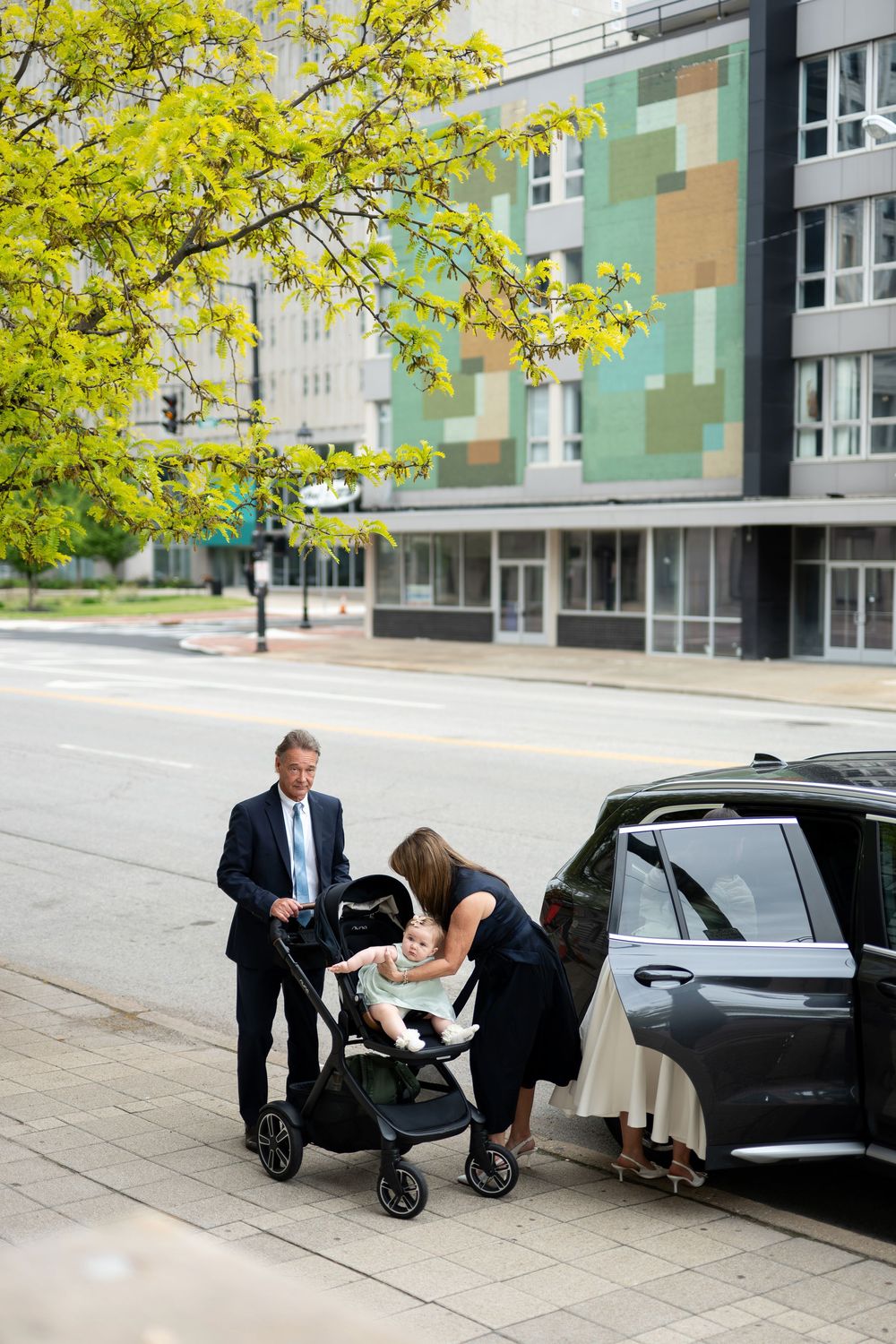 Family with baby in stroller beside an SUV on a city sidewalk during a wedding day outing.