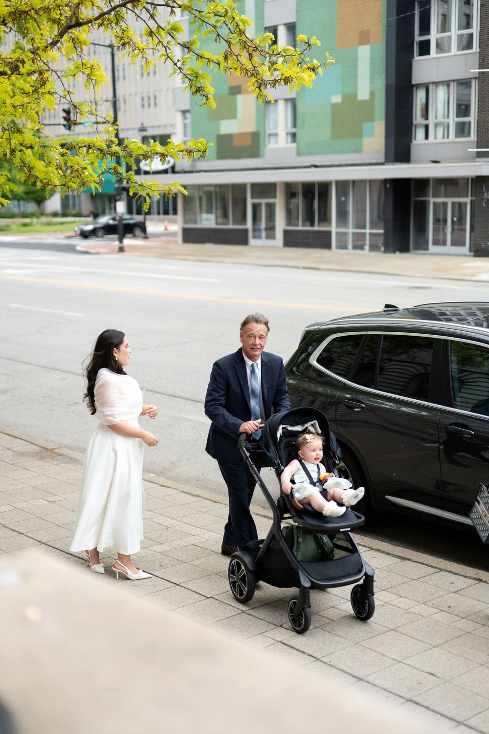 Bride in white dress and older man push baby stroller along urban sidewalk after courthouse wedding.