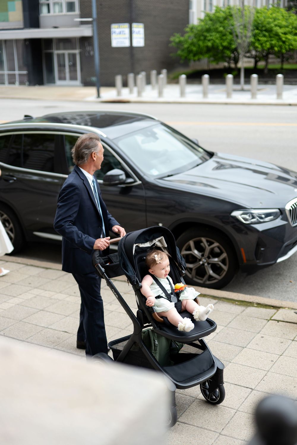 Grandfather in navy suit pushing baby in modern stroller on city sidewalk near black BMW SUV.