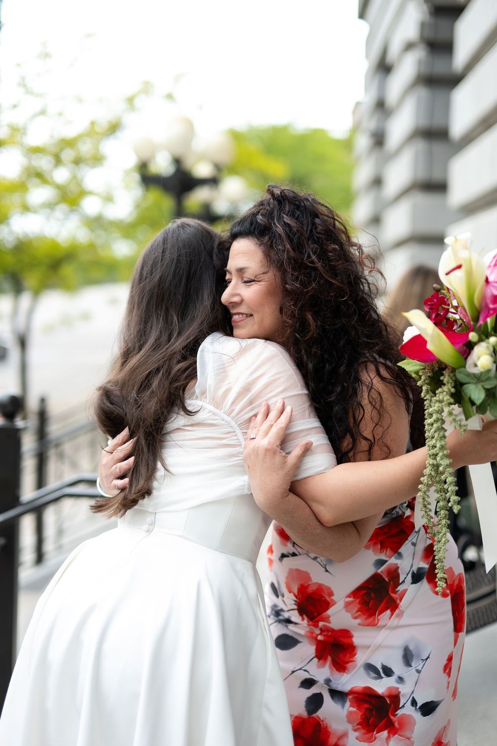 Two women embrace warmly outside a wedding venue, one in a white bridal gown and the other in a floral dress holding a bouquet.