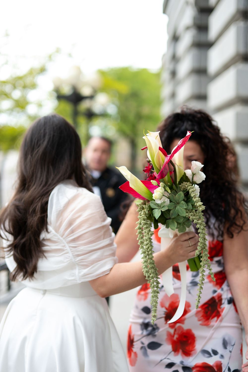 Bride in white dress receives floral bouquet with calla lilies and roses from a guest outdoors.