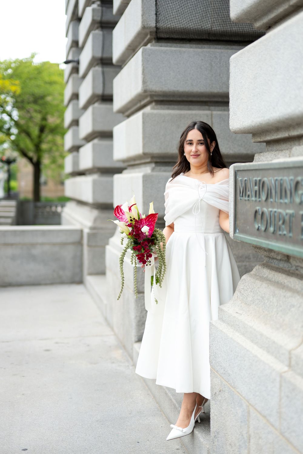 Bride in white off-shoulder midi dress holds vibrant red bouquet outside Mahoning County Courthouse.