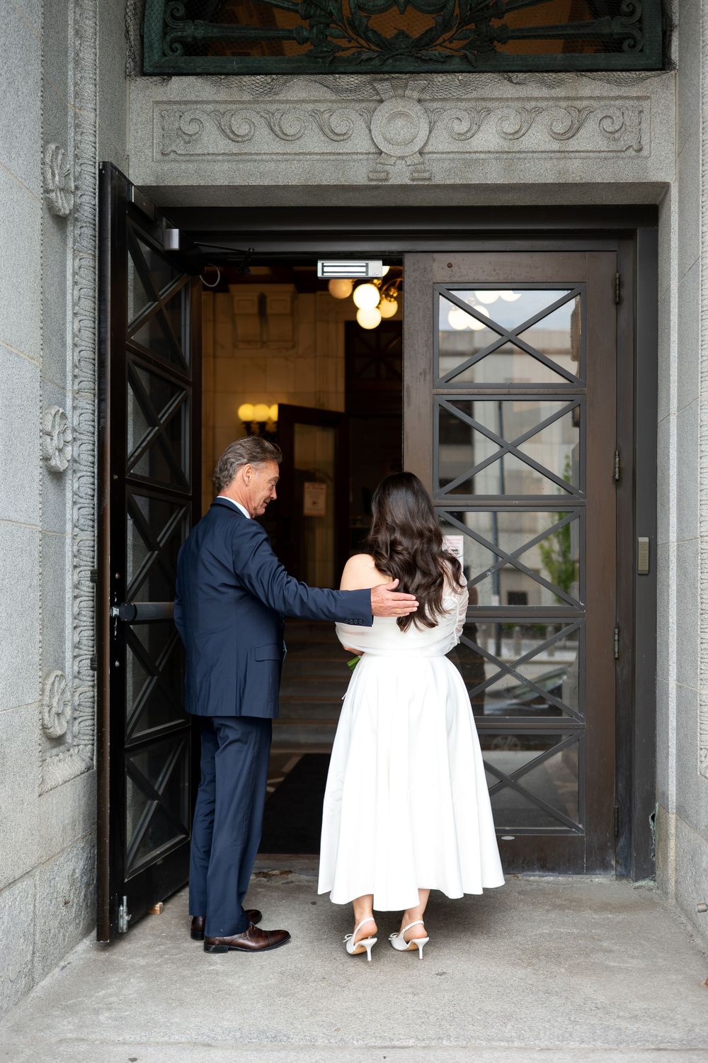 Groom in navy suit holding door open for bride in white midi dress at elegant stone building entrance.