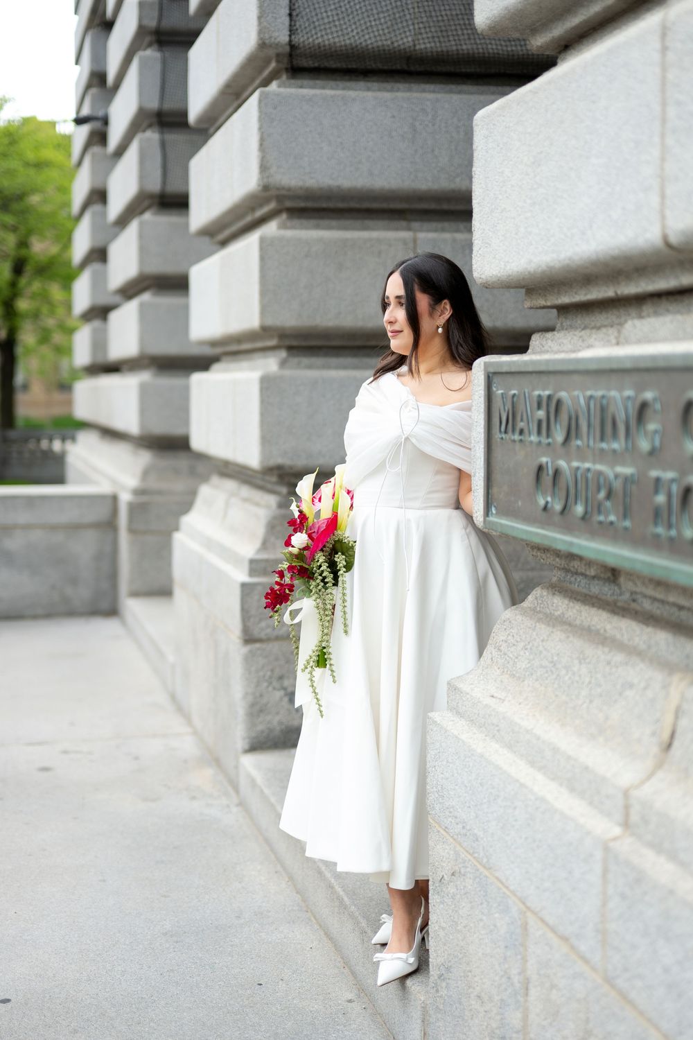 Bride in white off-shoulder midi dress holding colorful bouquet posing by Mahoning County courthouse stone wall.