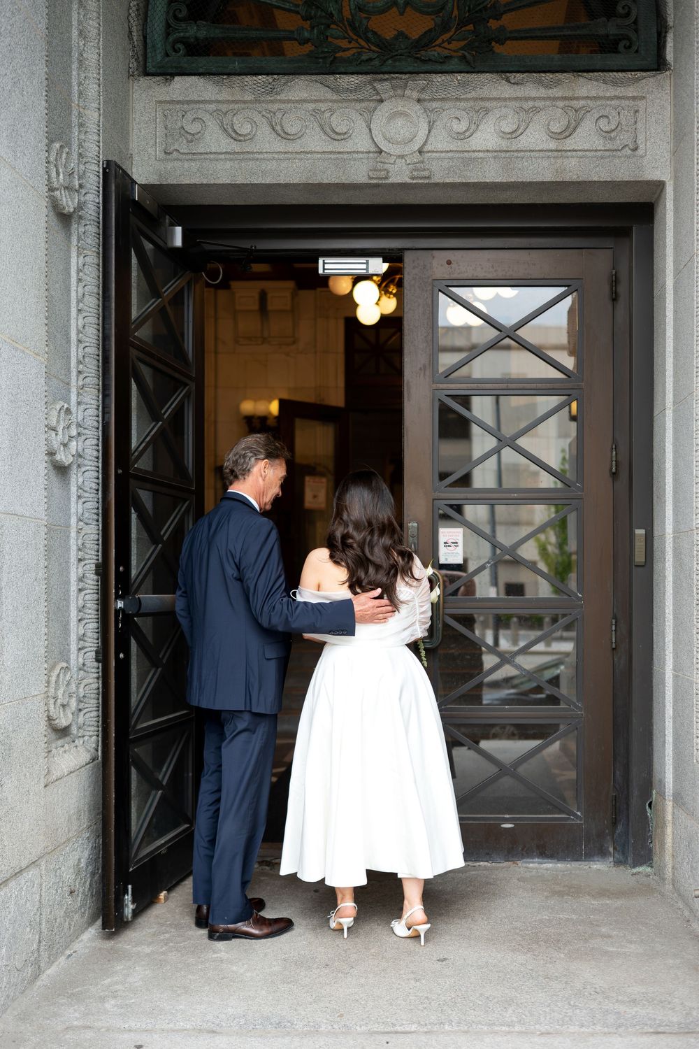 Couple in formal wedding attire entering through ornate double doors of a historic stone building.