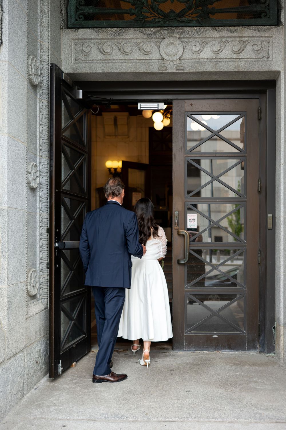 Bride in white midi dress and groom in navy suit entering ornate historic building through decorative doors.