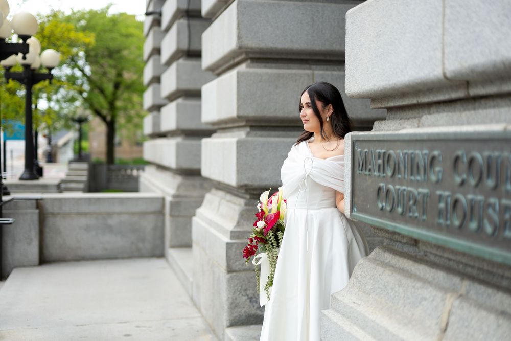 Elegant bride in white gown with wrap holds red bouquet leaning against Mahoning County Courthouse wall.