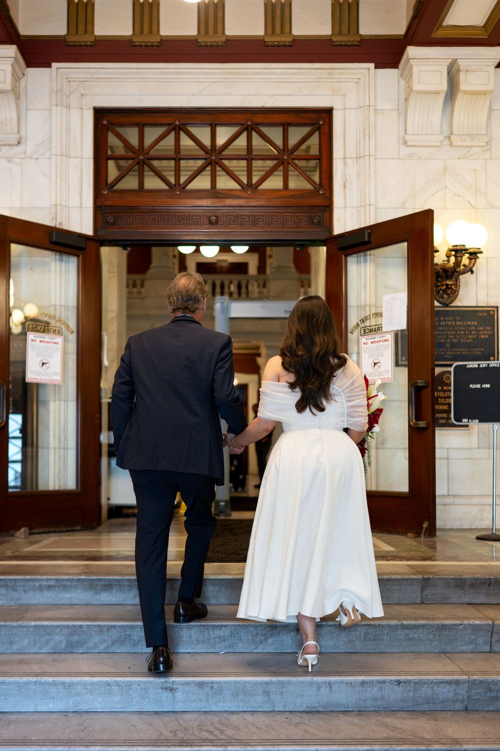 Bride in white off-shoulder gown and groom in dark suit enter ornate courthouse doors together.