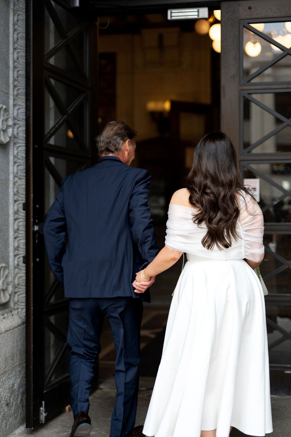Bride in white off-shoulder dress and groom in navy suit hold hands entering elegant building doors.